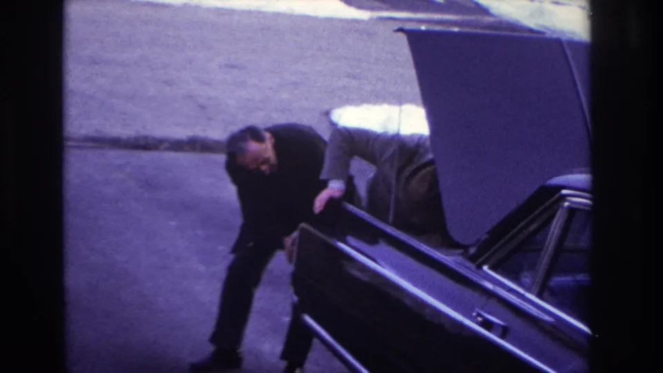 1966 Hoboken home-movie frame showing two suited men close to the open trunk area of a black car on a paved surface.