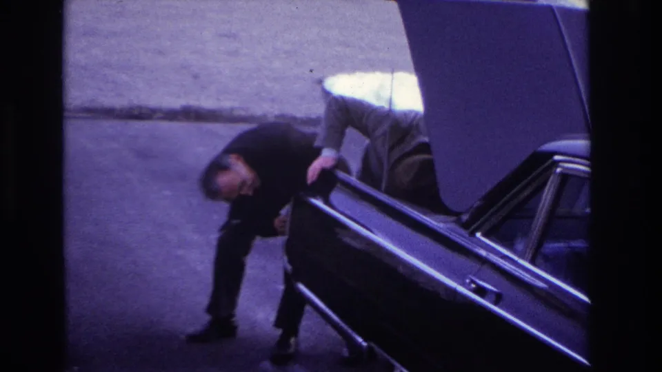 Final 1966 Hoboken frame with two suited men gathered at the open trunk of a black car, seen from an elevated home-movie viewpoint.