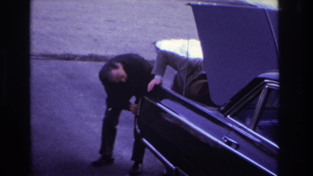 1966 Hoboken home-movie frame showing a man in a dark suit bending near the open trunk of a dark sedan on a quiet street.