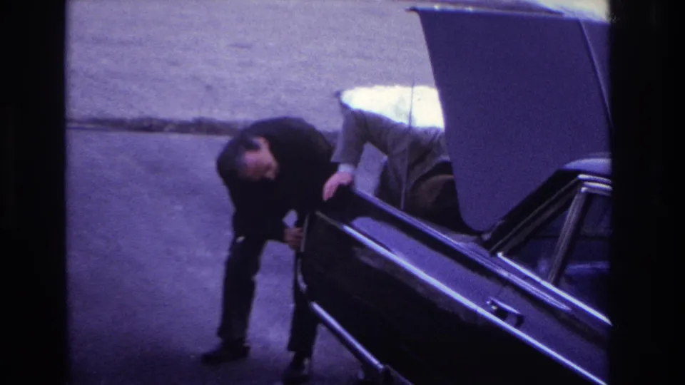 1966 Hoboken home-movie frame showing a man in a dark suit bending near the open trunk of a dark sedan on a quiet street.