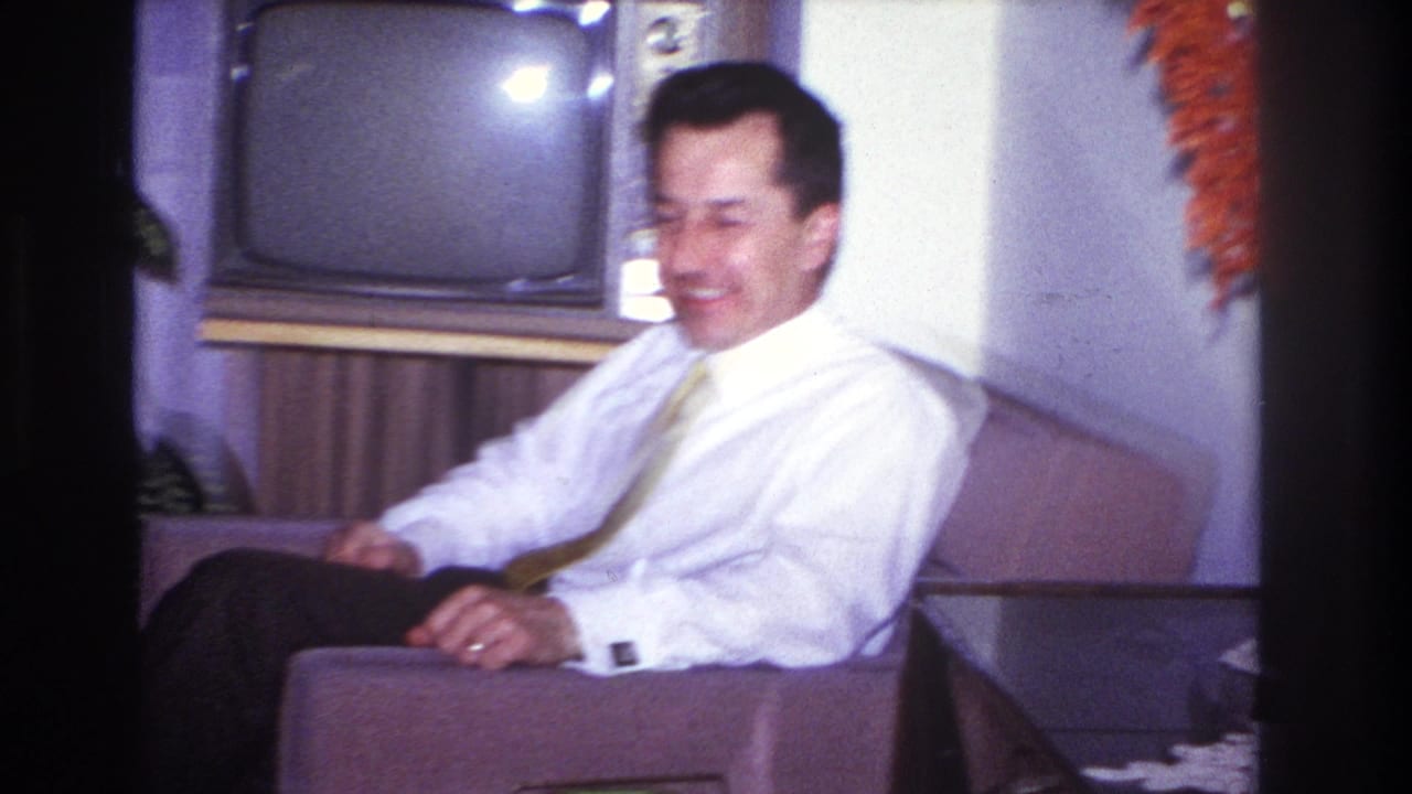 Man in white shirt and yellow tie seated in an armchair beside a vintage television.