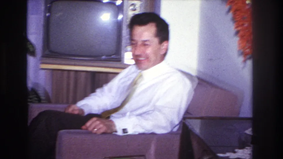 1966 Hoboken home-movie frame showing a man seated indoors in a mid-century chair near a vintage television set.