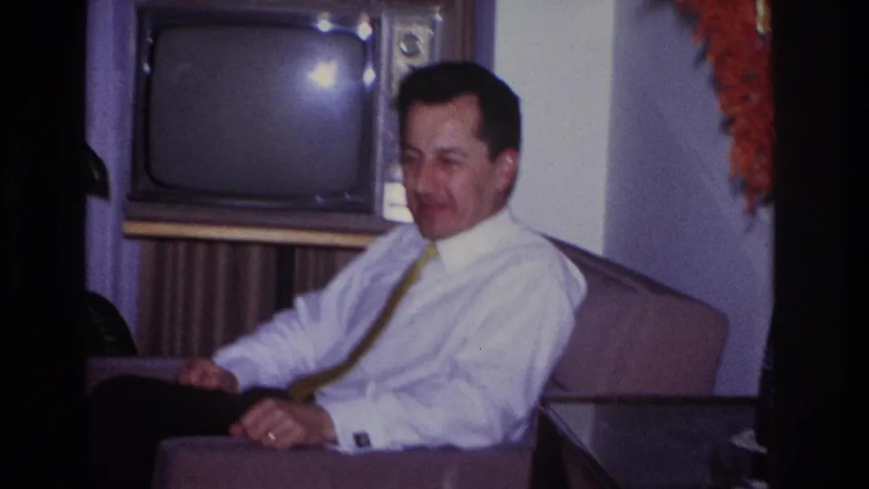 1966 Hoboken home-movie frame showing the seated man in profile near a vintage television and living-room furniture.
