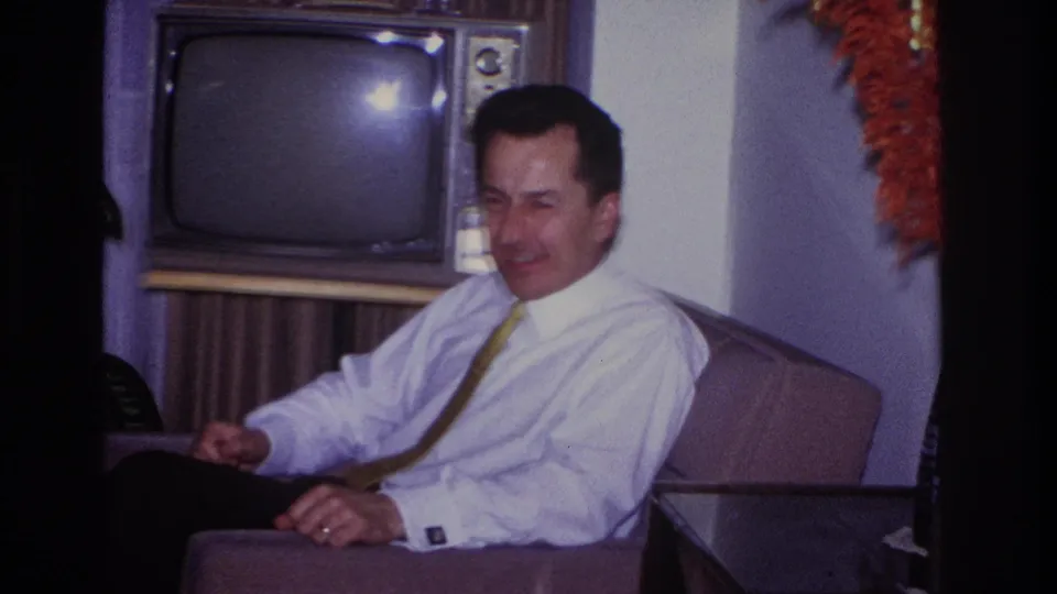 1966 Hoboken home-movie frame showing a smiling man in a white shirt and tie seated near a vintage television.
