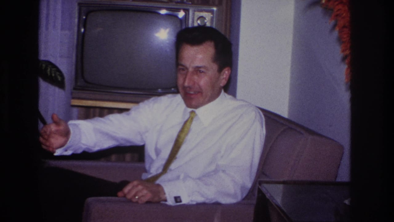 In a 1966 Hoboken home interior, a seated man in a white shirt and yellow tie smiles and gestures beside a wood-cased television.