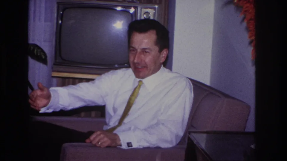 In a 1966 Hoboken home interior, a seated man in a white shirt and yellow tie smiles and gestures beside a wood-cased television.