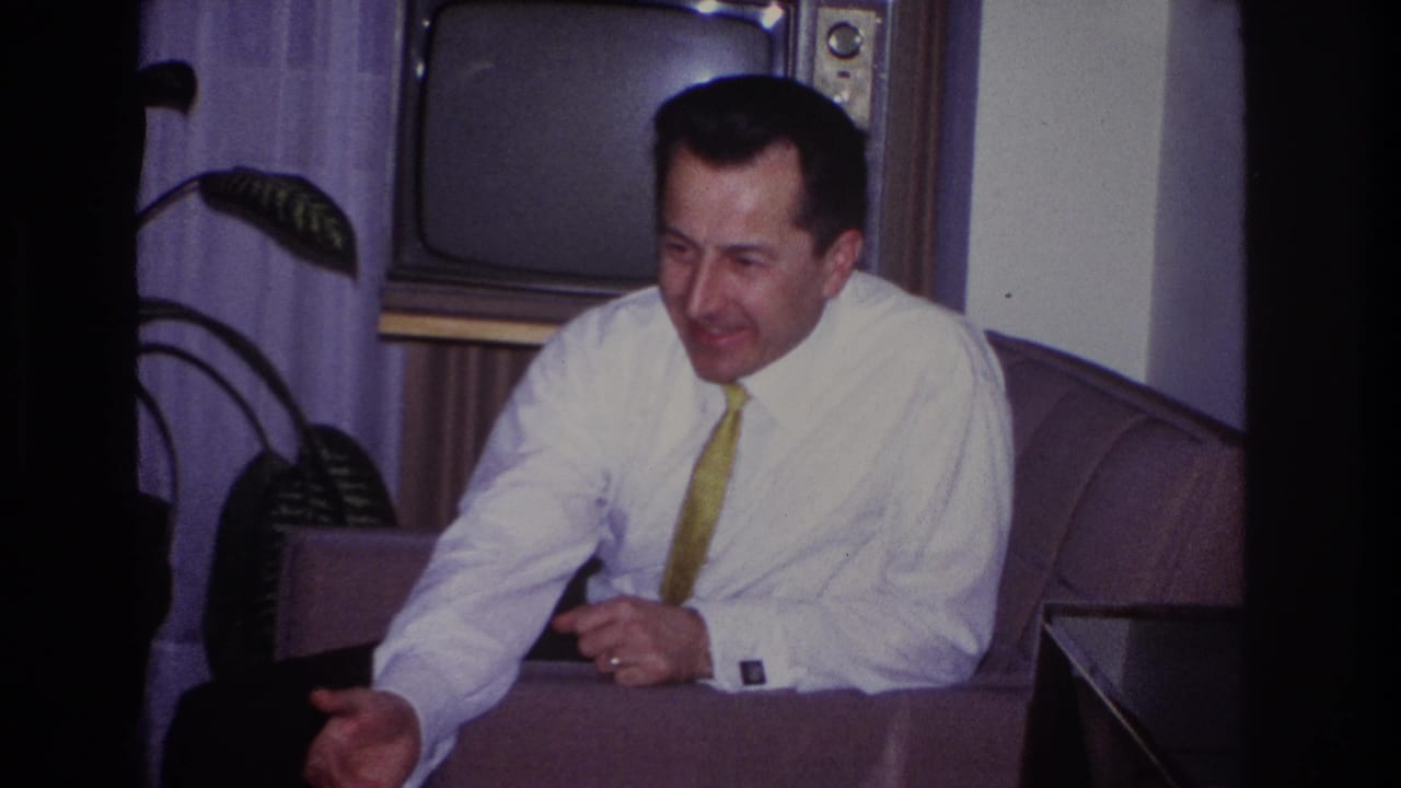 Man leans forward in an armchair with one hand extended, with a television and plant behind him.