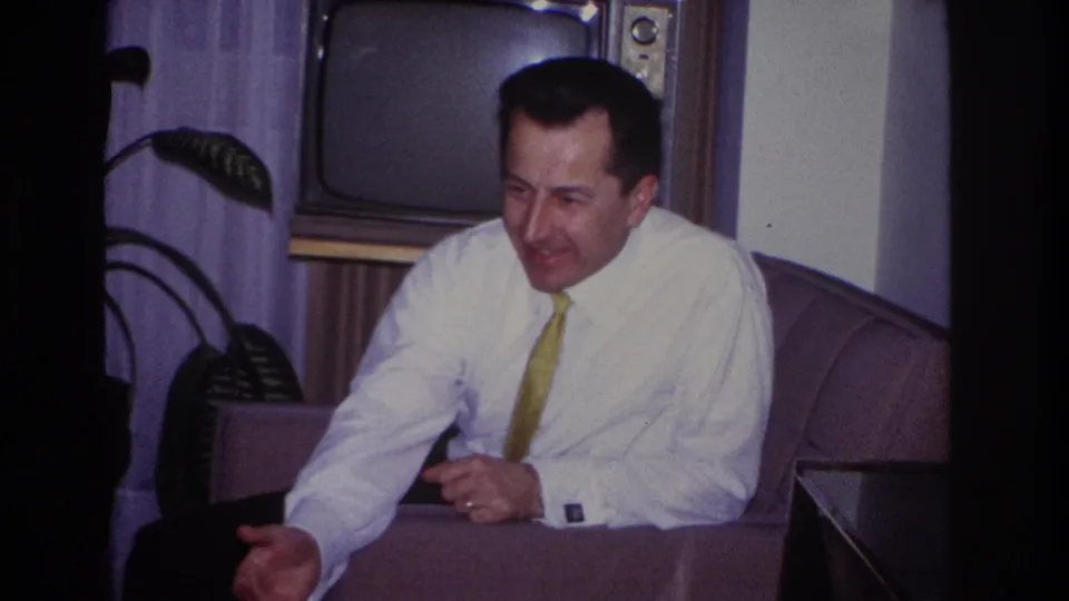 The man leans forward in a Hoboken living room, gesturing mid-conversation with a vintage television and plant in the background.