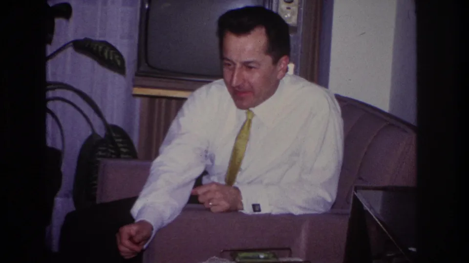A seated man in a white shirt and tie looks down briefly in the 1966 Hoboken living room, framed by curtains, a plant, and a television.