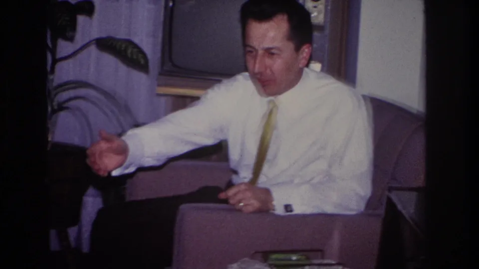 In the Hoboken home interior, the man extends his hand while speaking, with a wood-cased television and leafy plant behind him.