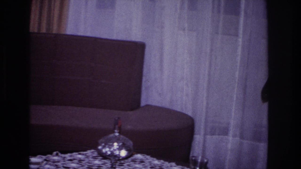 Dim living room with dark sofa, sheer curtains, glass table, and small decorative object.