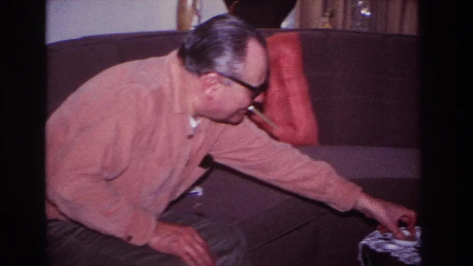 Older man in glasses and a pink cardigan leans from a Hoboken living-room sofa toward a lace-covered table in 1966 home-movie footage.