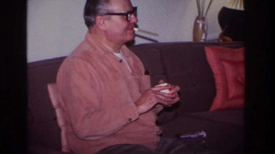 Older man in a pink cardigan sits upright on a dark sofa in a 1966 Hoboken interior, holding a cigarette and a small white item.