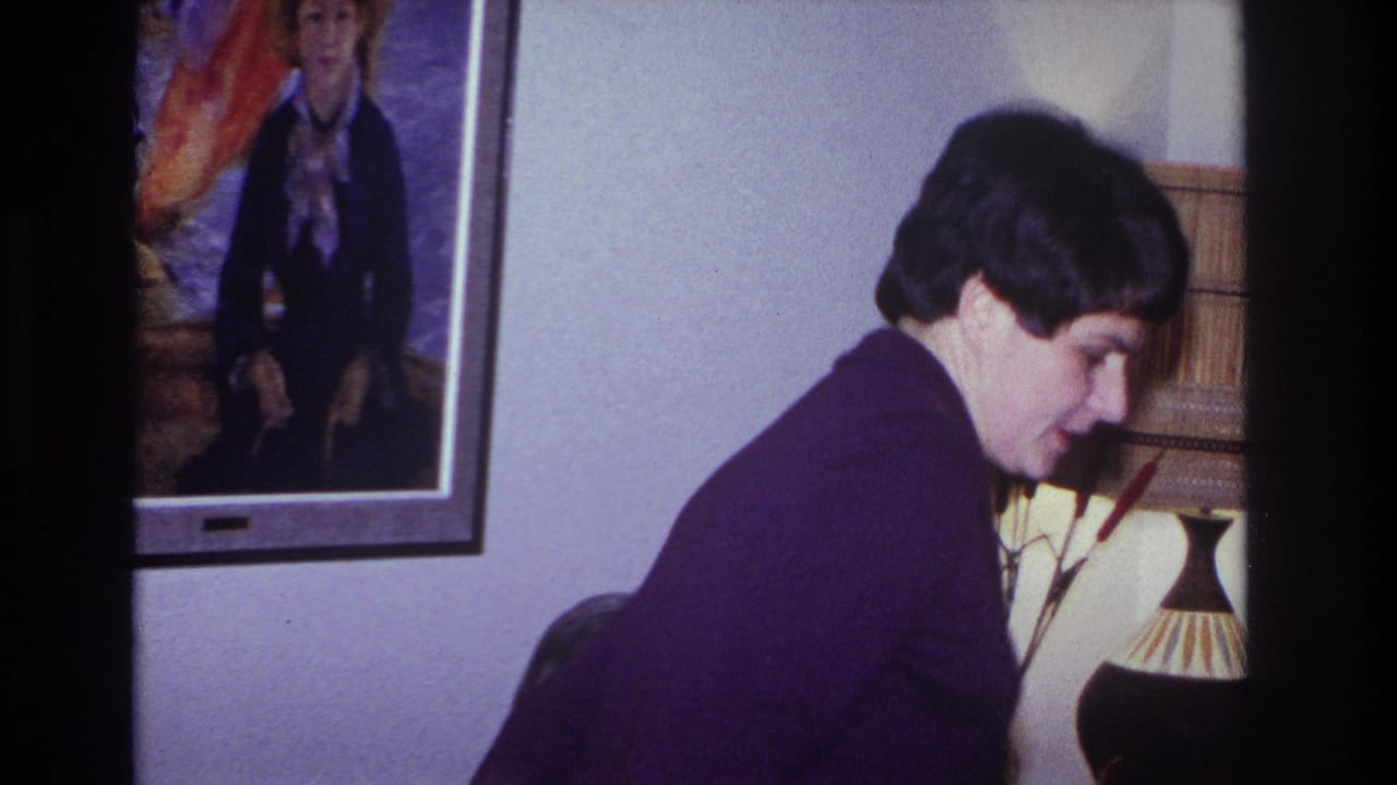In a 1966 Hoboken home-movie frame, a woman in a purple sweater leans near a living-room couch below framed artwork and a lamp.