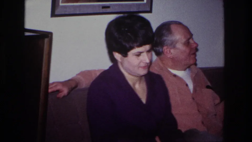 The woman sits beside the older man on a couch, with framed artwork and a living-room wall visible behind them.