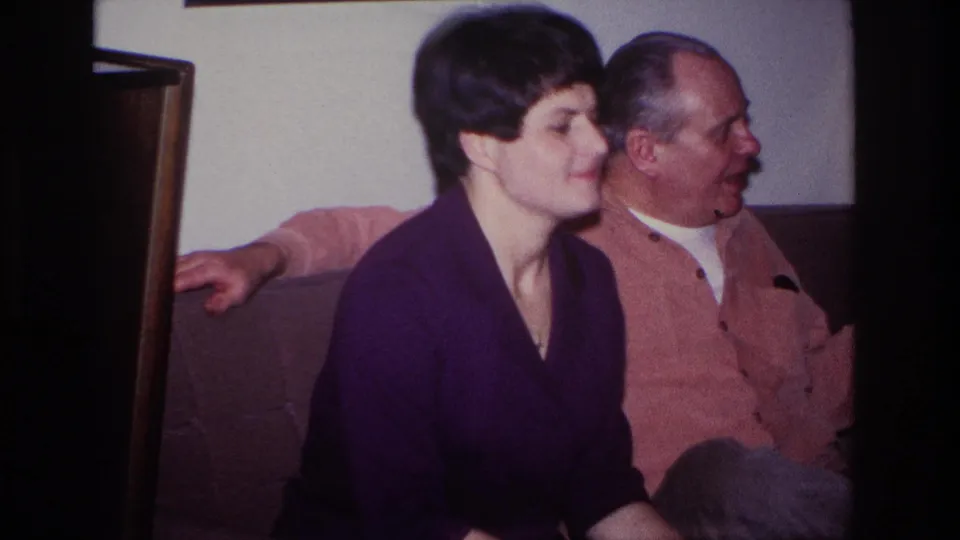 In the living room, the woman in purple faces forward while the older man in a pink cardigan turns slightly beside her.
