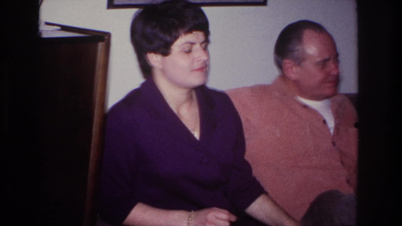 1966 Hoboken home-movie frame of a woman in a purple dress seated beside a man in a pink shirt on a couch.