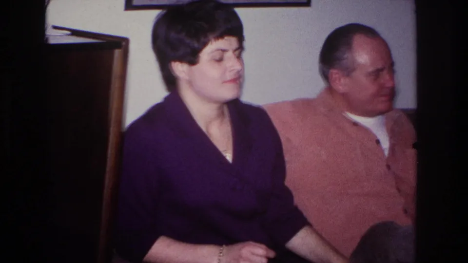 1966 Hoboken home-movie frame of a woman in a purple dress seated beside a man in a pink shirt on a couch.