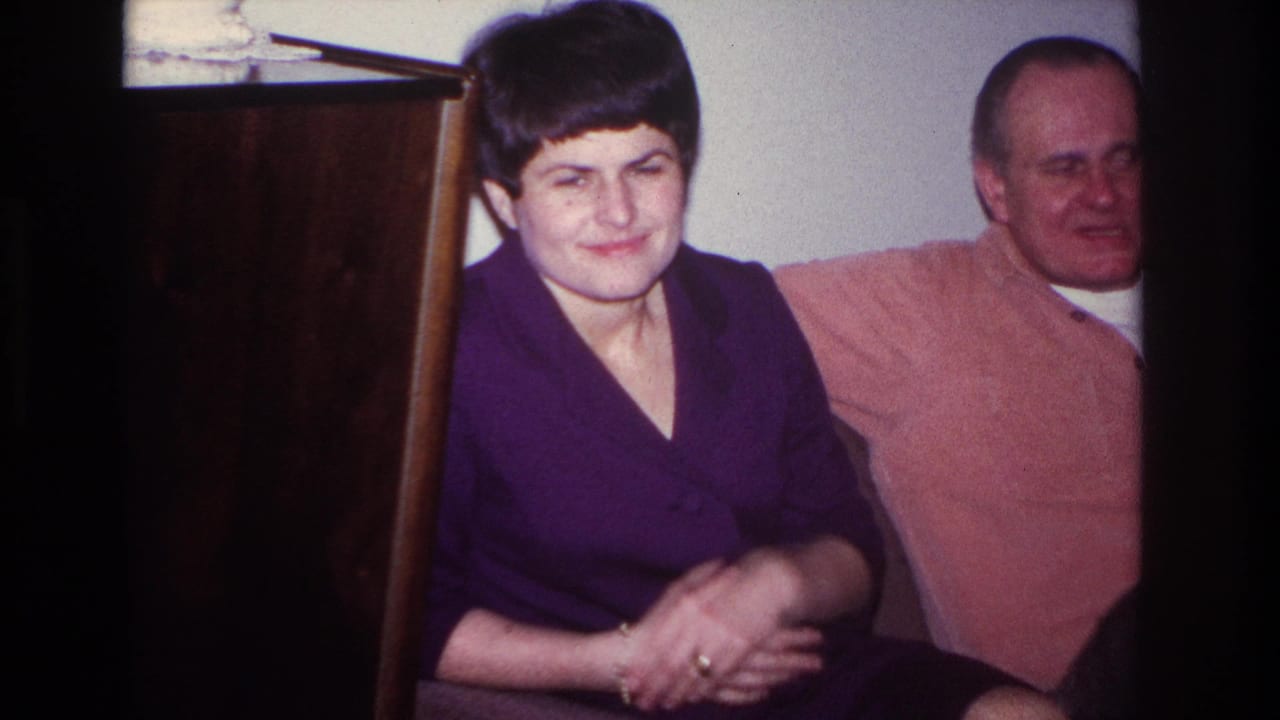 Woman smiles toward the camera while a seated man is partly visible beside her.