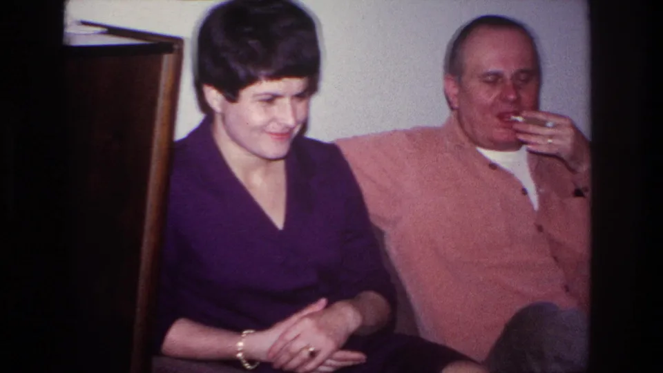 1966 Hoboken home-movie frame of a woman smiling downward while a man beside her smokes on the couch.
