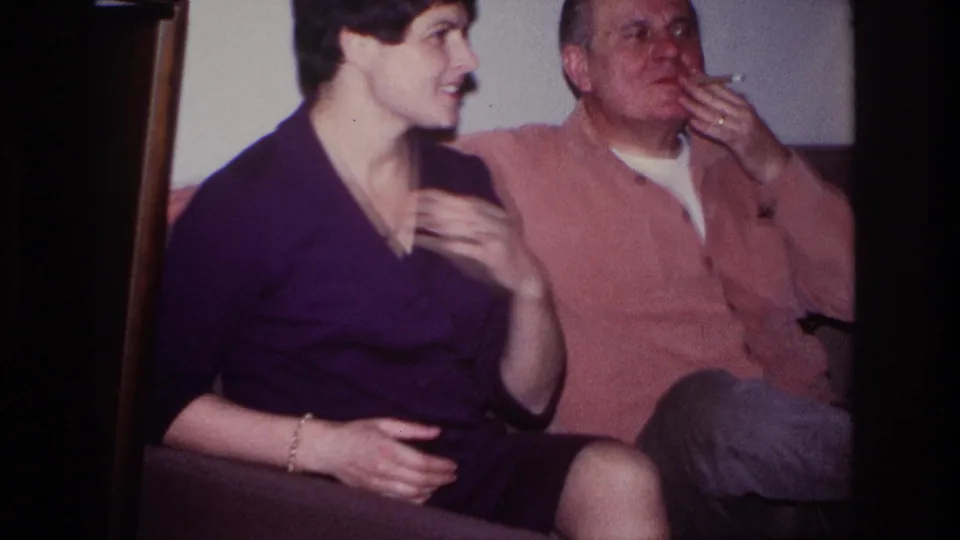 1966 Hoboken home-movie frame showing the woman turned toward the man as he smokes indoors on the couch.