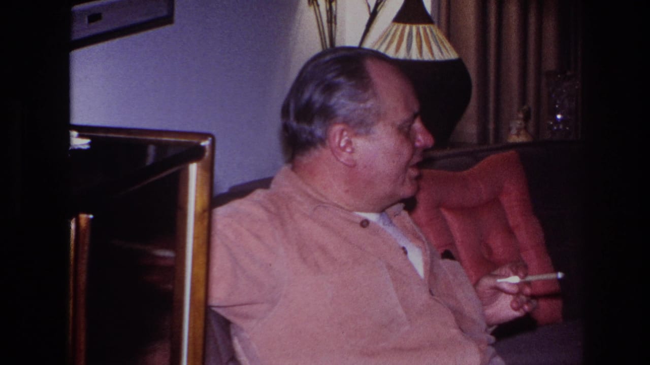 The older man remains seated in profile in the Hoboken living room, with curtains and a pleated lampshade behind him.