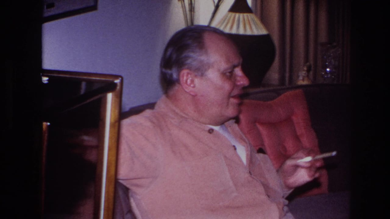 The man looks sideways while seated in the Hoboken living room, with a red chair, lamp and curtains visible around him.