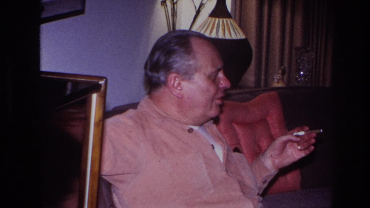 In a 1966 Hoboken home-movie frame, a middle-aged man in a pink button-up shirt sits in a red armchair and turns to the side while holding a cigarette.