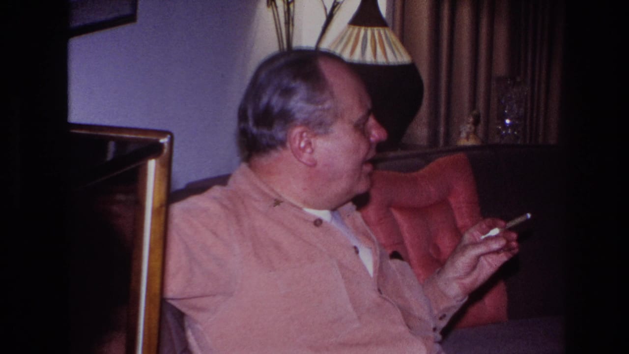 Man turns toward the right while holding a cigarette near a lamp and curtains.