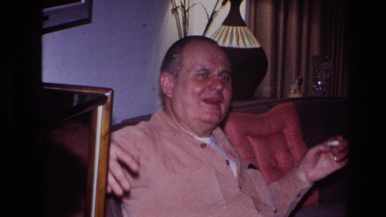 The seated man faces more toward the camera, appearing mid-story or mid-laugh as he gestures in the living-room chair with the cigarette still in hand.