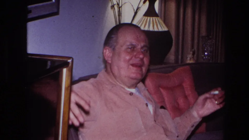 The seated man faces more toward the camera, appearing mid-story or mid-laugh as he gestures in the living-room chair with the cigarette still in hand.