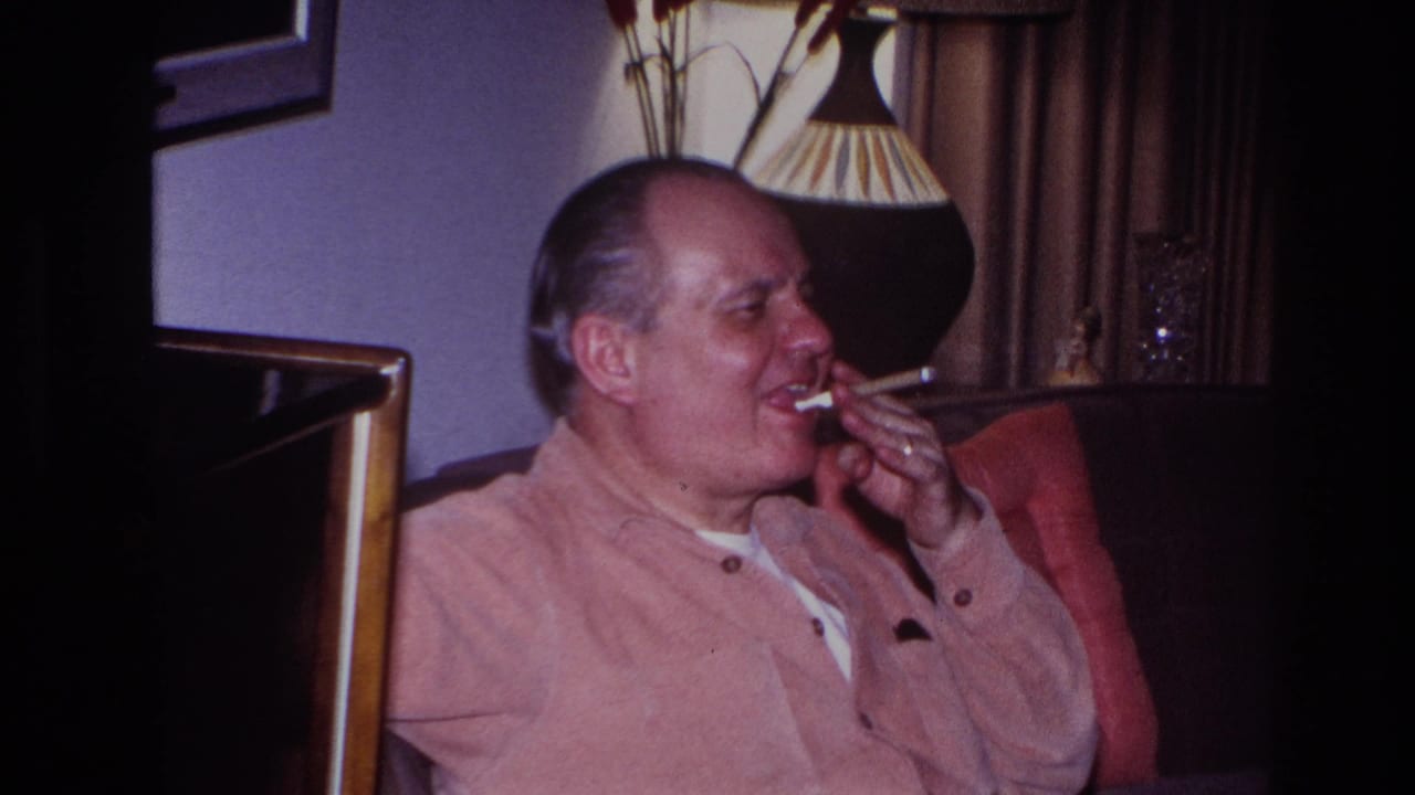 The man smiles or laughs with the cigarette near his mouth, seated in the red armchair against a dim domestic background of lamp, curtains, and wall decor.