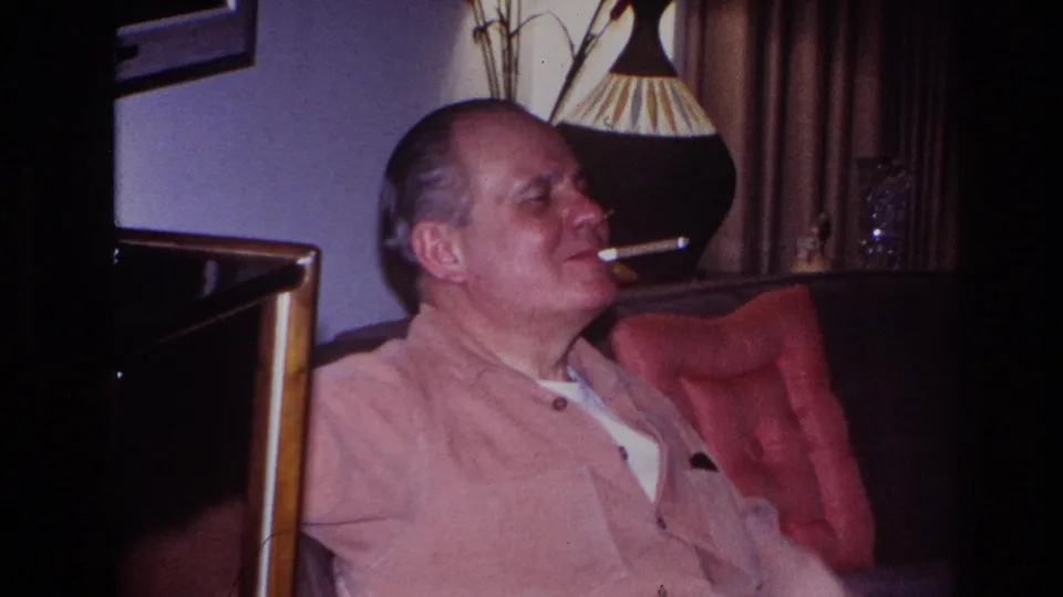 In 1966 Hoboken home-movie footage, a man in a light pink shirt sits on a dark sofa smoking beside a lamp and curtains.