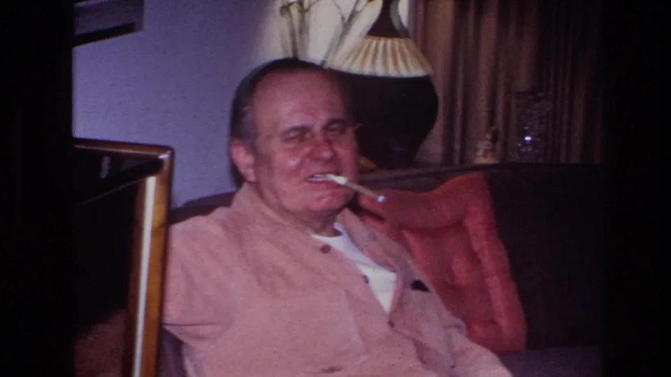 A man in a light shirt reclines on a dark sofa in a Hoboken living room, smoking as curtains and a lamp sit behind him.