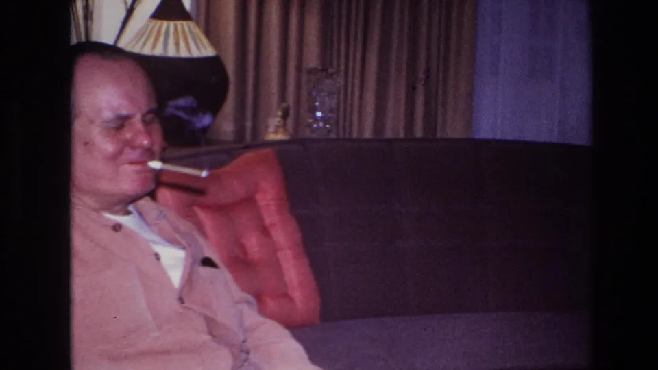 The home-movie frame shows the man partly cropped at left, still smoking on the sofa with a lamp, curtains, and cushion visible.
