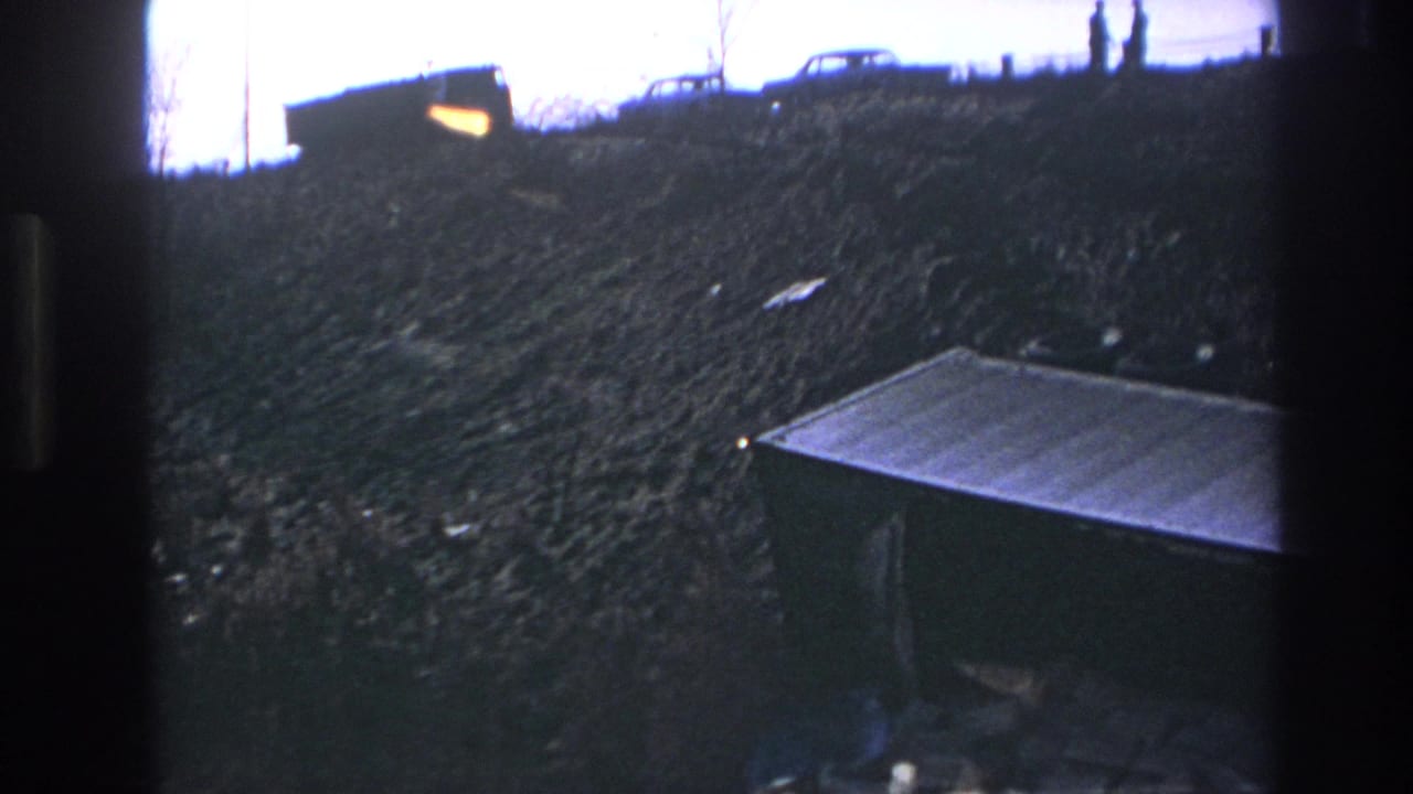 1966 Hoboken 8mm frame showing an overgrown slope, ridge-top cars, distant figures, and a dark metal container in the foreground.
