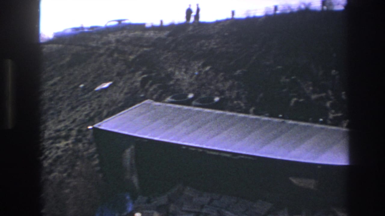 Distant figures on a ridge above a sloped debris area and metal container.