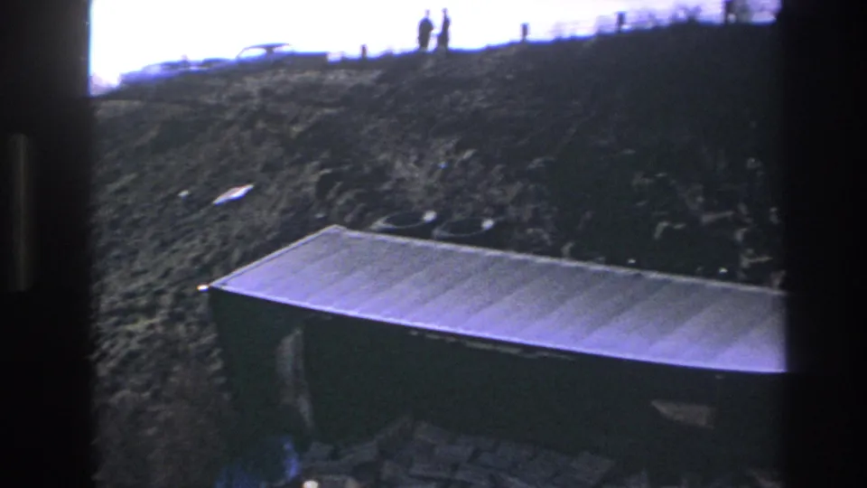 Hoboken 1966 home-movie frame with silhouetted people on a ridge, cars nearby, and a large metal container below.