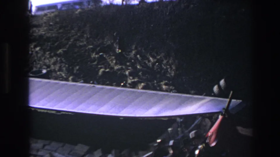 8mm frame from Hoboken showing the edge of a large metal container, weeds, debris, and partial foreground objects in low light.