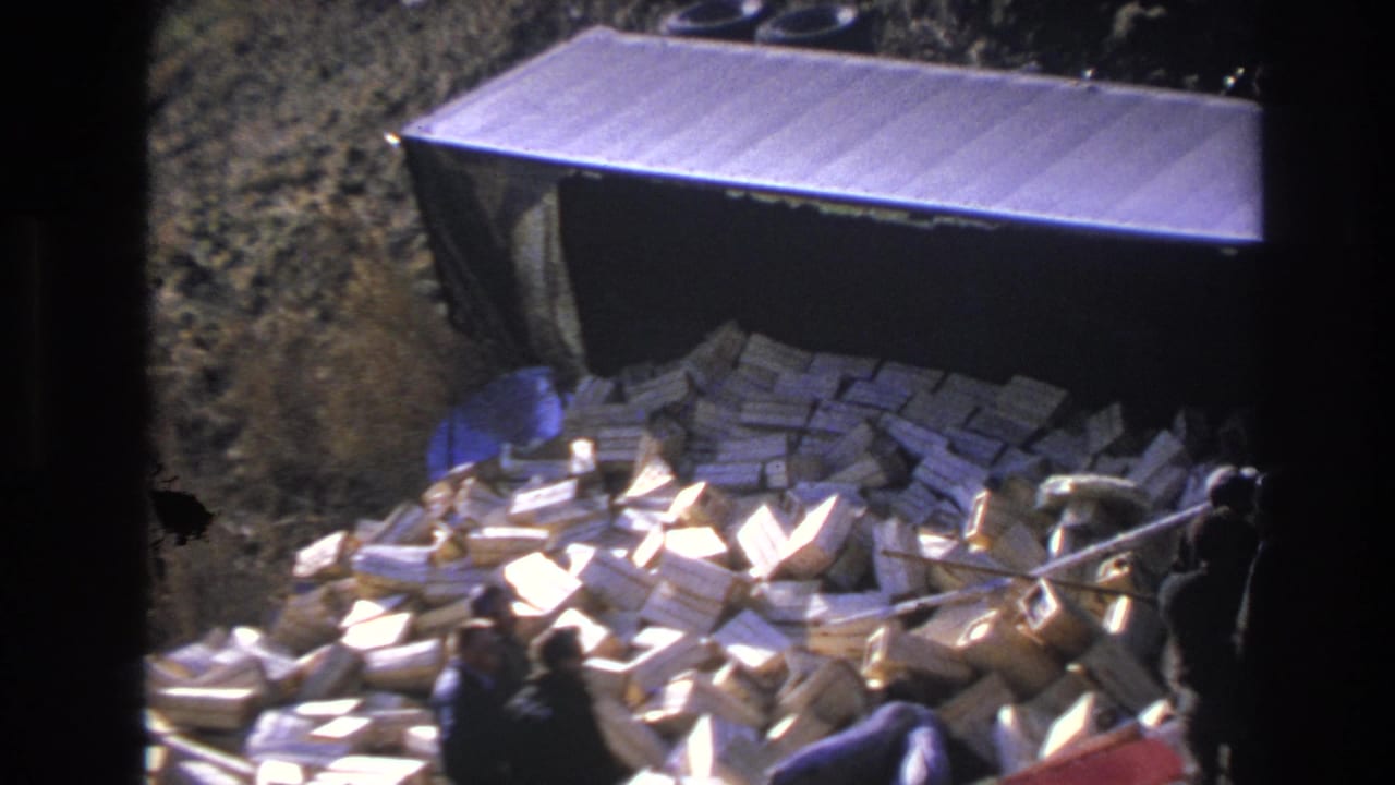 Hoboken, New Jersey 1966 home-movie frame showing men gathered near spilled boxes and debris below a dark truck body on a rough grassy slope.