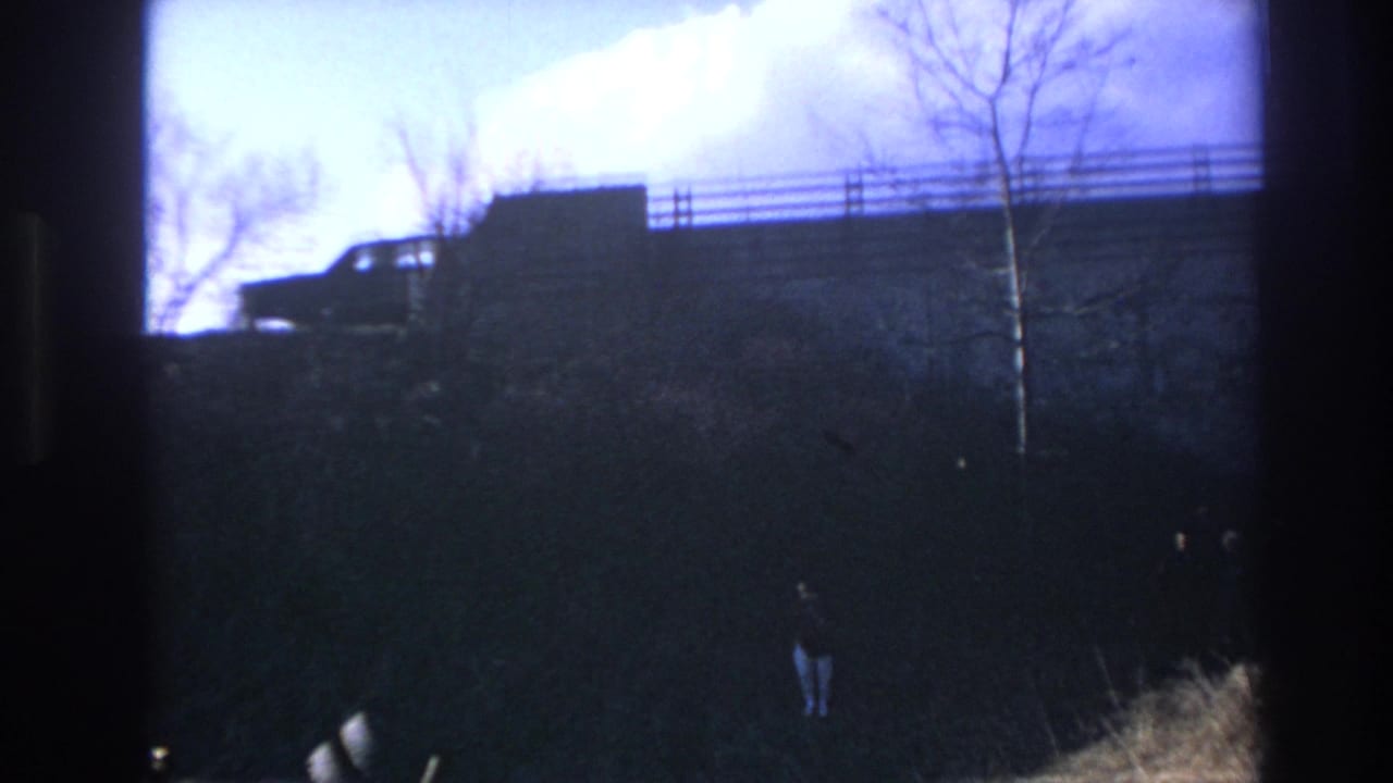 1966 Hoboken home-movie frame showing a dark car on an elevated roadway, bare trees, and two small figures on the embankment below.