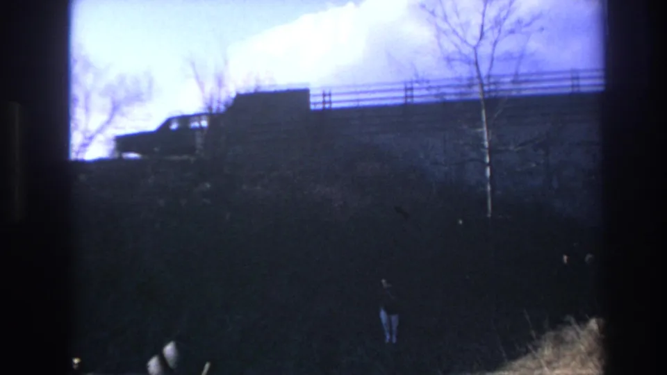 1966 Hoboken home-movie frame showing a dark car on an elevated roadway, bare trees, and two small figures on the embankment below.