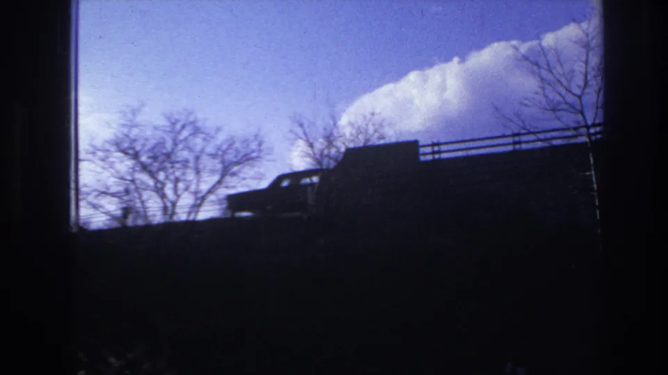 1966 Hoboken frame with a dark car on the raised road, leafless trees, railings, and a bright clouded sky.
