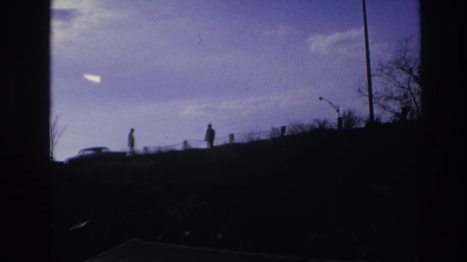 1966 Hoboken frame showing two distant silhouetted people on the embankment, a car at left, bare trees, and a lamppost against the sky.