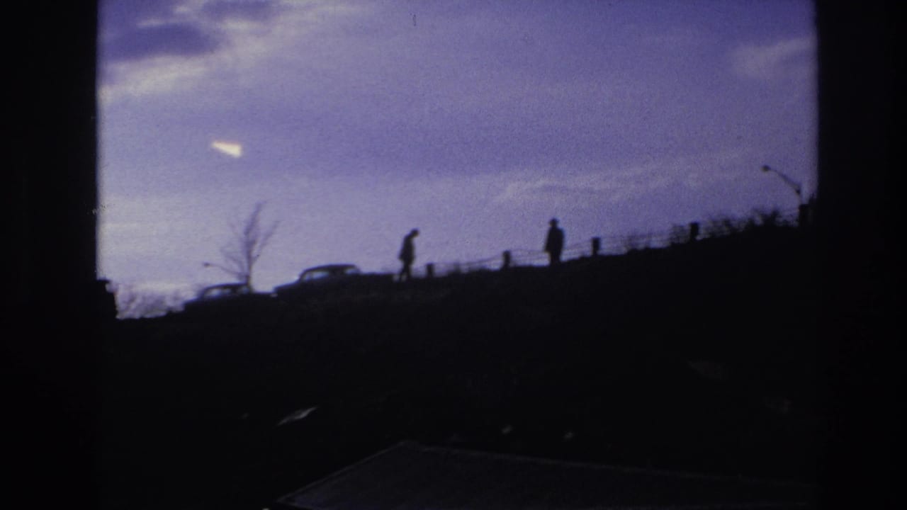 1966 Hoboken-area 8mm frame showing silhouetted figures and cars on a ridge, seen through dark foreground obstruction under a purple sky.