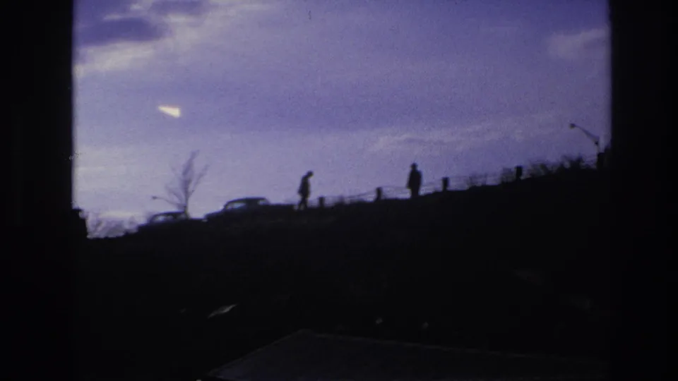 1966 Hoboken-area 8mm frame showing silhouetted figures and cars on a ridge, seen through dark foreground obstruction under a purple sky.