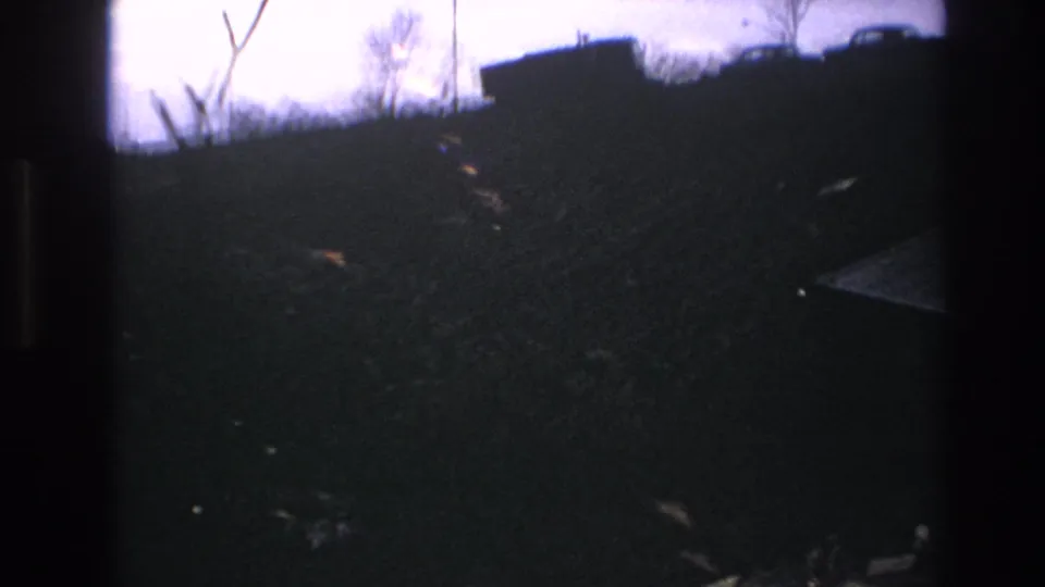 1966 Hoboken-area 8mm frame showing a dim, blurred slope with brush, bare branches, and a vehicle silhouette near the top edge.