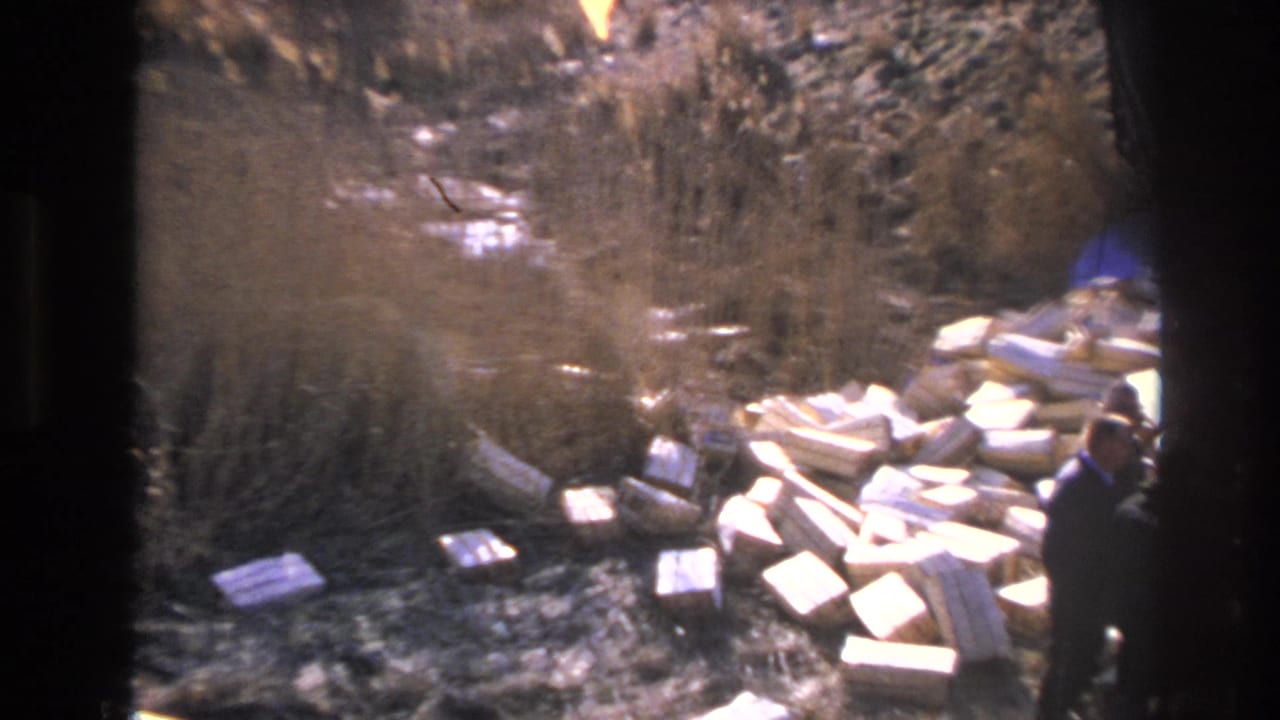 1966 Hoboken home-movie frame showing men near a pile of white blocks beside dry grass and a dirt embankment.