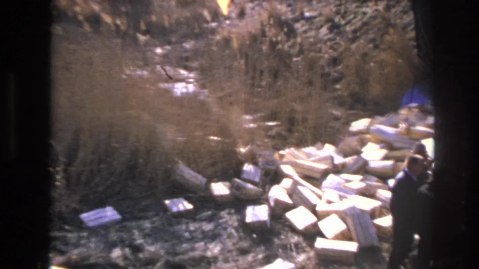 1966 Hoboken home-movie frame showing men near a pile of white blocks beside dry grass and a dirt embankment.