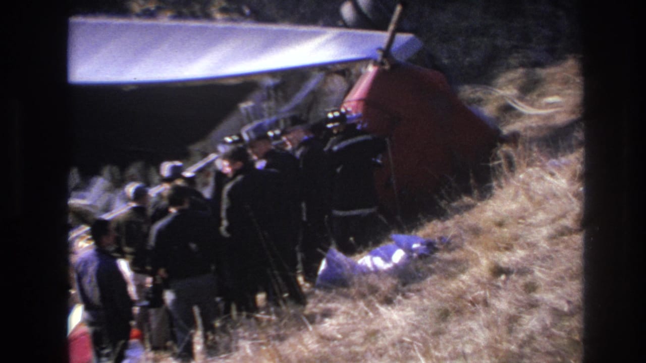 A group of men stands close to the overturned aircraft, with dry grass and debris surrounding the crash site.